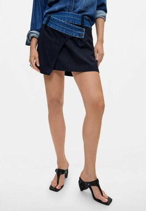 Mujer que lleva una falda corta de rayas finas con cinturilla de denim en capas y sandalias negras de tacón con punta cuadrada.