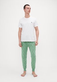 T-shirt en coton blanc avec un petit logo noir sur la poitrine, associé à un pantalon de pyjama vert présentant un motif de chevaux blancs répétitif.