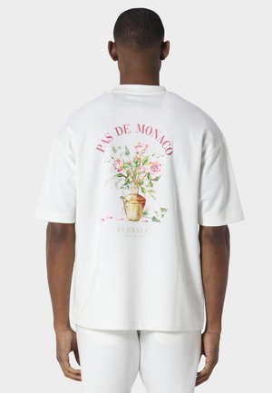 Homme portant une chemise blanche et un pantalon, le dos de la chemise montre une illustration d’un vase floral avec le texte « PAS DE MONACO » et « FLORALE ».