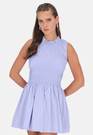 Młoda kobieta o długich włosach, ubrana w lekko fioletową sukienkę na ramiączkach z marszczonym gorsem i rozkloszowanym dołem, stojąca na tle gładkiego tła.