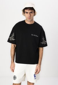 Άνδρας που φοράει μαύρο φαρδύ t-shirt με λευκή κεντητή ανθοδέσμη στο μανίκι και λευκό σορτς, στέκεται μπροστά σε λευκό απλό φόντο.