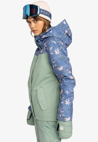 Ski jacket with a floral blue upper and solid green lower. Features a hood, side pockets, and matching green mitts, made of textured fabric.