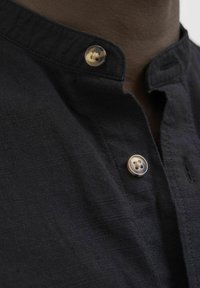 Camicia nera di lino con un piccolo colletto a righe, caratterizzata da un'importante bottone dorato con un motivo a croce e una superficie di tessuto testurizzata.