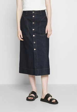 Denim midi-rok, donkerblauw met zichtbare stiksels, sluiting met knopen aan de voorkant, twee grote voorkruiden, gecombineerd met zwarte sandalen.
