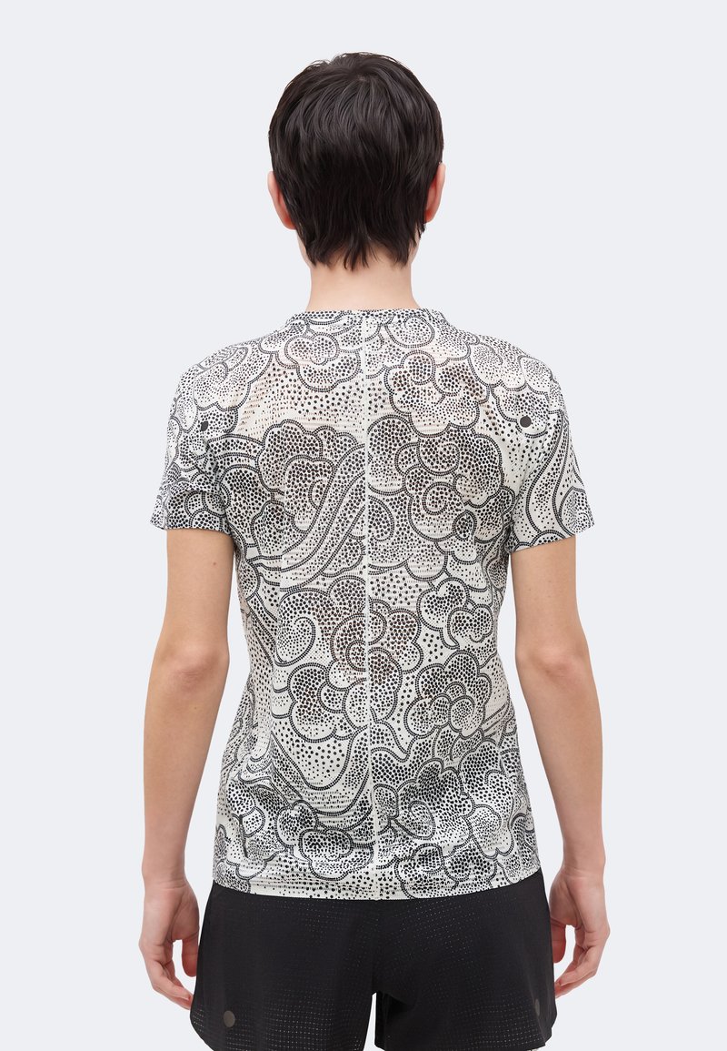 Person, die mit dem Rücken zur Kamera steht, trägt ein weißes kurzärmeliges Hemd mit einem schwarzen abstrakten Blumen- und Punktmuster, kombiniert mit schwarzen Shorts.