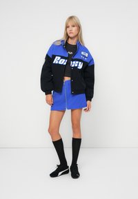 Blauwe en zwarte jas met "Roddy" grafiek, gecombineerd met een blauwe ritsskort. Zwarte kniehoge sokken en zwarte sneakers maken de look compleet.
