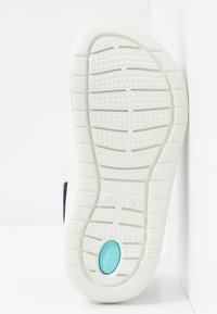 Semelle de chaussure blanche avec motif de grip texturé et un petit détail ovale turquoise près du talon.