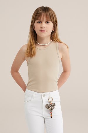 Lány egyenes világosbarna hajjal bézs ujjatlan felsőt, fehér nadrágot visel leopárdmintás szív alakú kulcstartóval és egy fehér gyöngyös nyaklánccal.