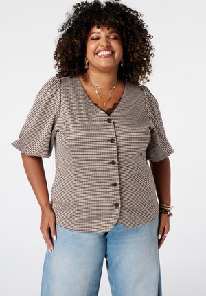 Vrouw met krullend haar glimlacht met gesloten ogen, draagt een geruit blouse met knopen en lichtblauwe spijkerbroek, staand tegen een witte achtergrond.