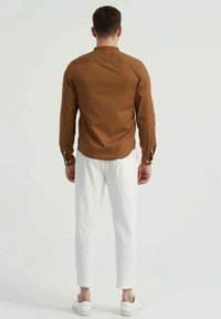 Chemise marron à manches longues avec un col mandarin, associée à un pantalon texturé blanc et des baskets blanches. La vue arrière met en évidence le design épuré de la chemise.
