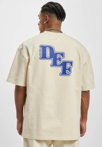 Light beige cotton t-shirt featuring large blue "DEF" graphic print on the back, short sleeves, relaxed fit, and a crew neck design.