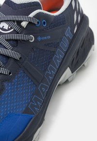 Marineblå atletiske sko med Gore-Tex, mesh-overdel, grå accenter, struktureret design og kontrasterende hvid sål; har et fremtrædende "MAMMUT"-brand.