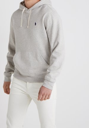 Person iført en lysegrå hættetrøje med en forlomme og hvide snore, kombineret med hvide bukser.