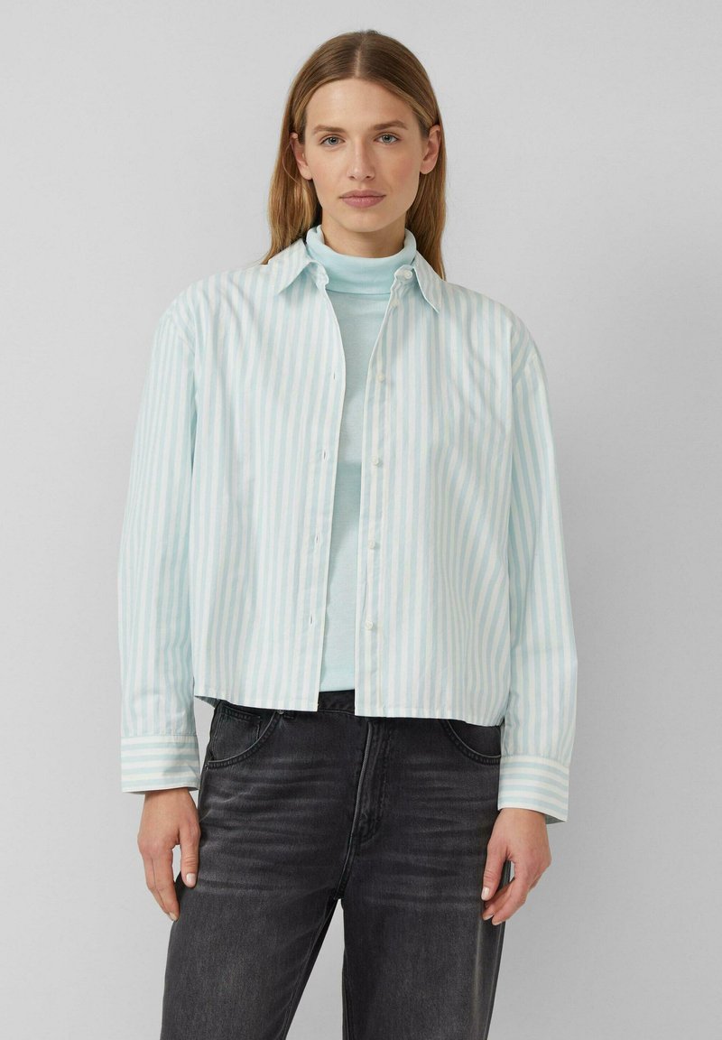 Vrouw met recht lichtbruin haar, gekleed in een lichtblauwe coltrui, een wit-blauw gestreept overhemd met knopen, en zwarte jeans.