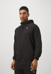 Schwarze leichte Jacke mit Kapuze, durchgehendem Reißverschluss vorne, elastischen Bündchen und einem weißen Logo auf der linken Brust. Glatte, matte Textur.