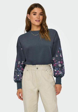 Dark gray sweatshirt with floral embroidery in pink, purple, and white on puffed sleeves; paired with high-waisted beige trousers.