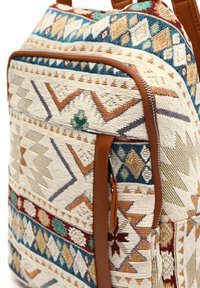 Textile backpack featuring intricate geometric patterns in cream, blue, and rust, with brown leather accents and a zippered pocket.