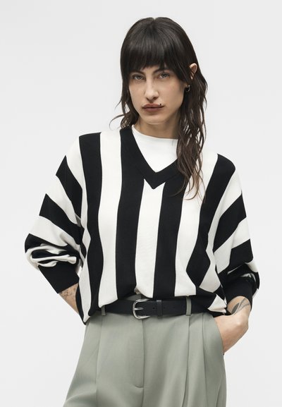 Άτομο με σκούρα μαλλιά, φοράει πουλόβερ με μαύρες και λευκές κάθετες ρίγες, ανοιχτό γκρι παντελόνι και μαύρη ζώνη, με τα χέρια στις τσέπες.