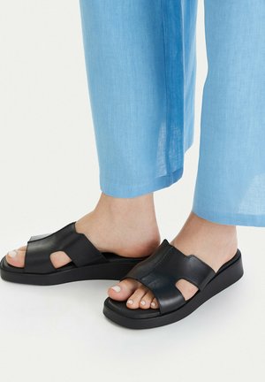 Pies con sandalias negras sin cordones con recortes combinadas con pantalones cortos azul claro sobre un fondo blanco.