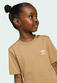 Smeđi pamučni majica s bijelim Adidas logotipom na lijevoj strani prsnog koša. Dizajn s okruglim izrezom i kratkim rukavima, glatke teksture i ležernog kroja.