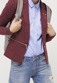 Blazer bordeaux con tasche con zip sopra una camicia azzurra a bottoni e jeans in denim strappati. Spalline dello zaino argentate visibili.