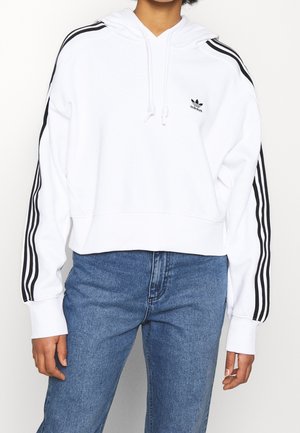 Witte crop hoodie met zwarte strepen op de mouwen, voorzien van een trekkoordkap en een klein Adidas-logo op de borst. Draag met blauwe jeans.