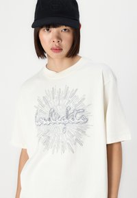 Cremefarvet oversized t-shirt med et metalisk sølvgrafikdesign, der fremhæver ordet "lowlights" og solstrålemønstre.