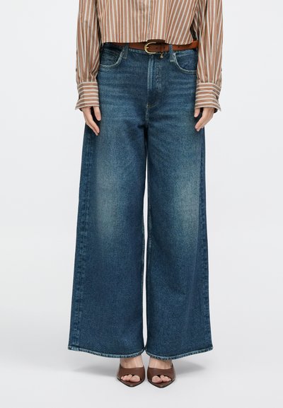 Laiade säärega sinised teksapüksid, millel on tuhmunud tekstuur, kõrge piht ja viie taskuga disain. Kanditud pruuni vöö ning pruunide kontsakingadega.