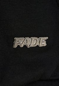 Czarny materiał o prążkowanej fakturze z metalicznym srebrnym logo z napisem "FADE" w pogrubionej, stylizowanej czcionce.
