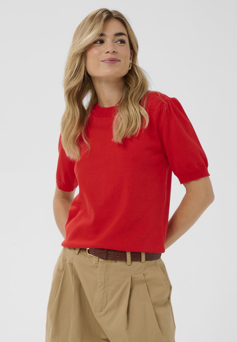 Punane lühikeste varrukatega särk ümmarguse kaelusega ja kergete volangidega varrukatega, kombineeritud beežide pükstega ja pruun vöö. Sile tekstuur.