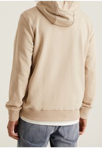 Uomo che indossa un hoodie beige e jeans blu, mostrato di spalle su uno sfondo neutro e uniforme.
