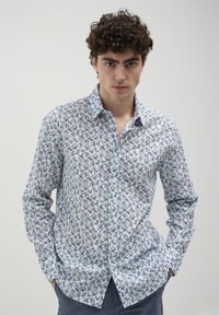 Giovane uomo con capelli ricci che indossa una camicia bianca con bottoni e motivi floreali blu, mani in tasca, di fronte a uno sfondo chiaro e semplice.