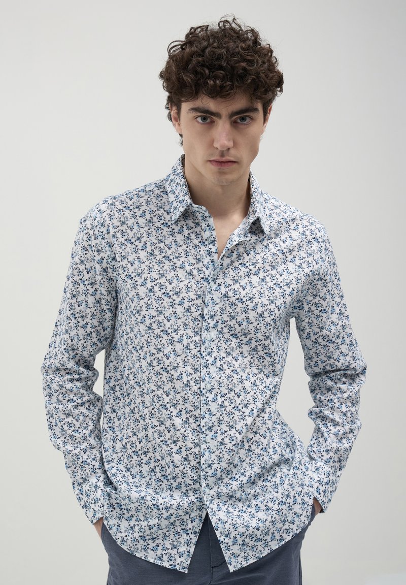 Giovane uomo con capelli ricci che indossa una camicia bianca con bottoni e motivi floreali blu, mani in tasca, di fronte a uno sfondo chiaro e semplice.