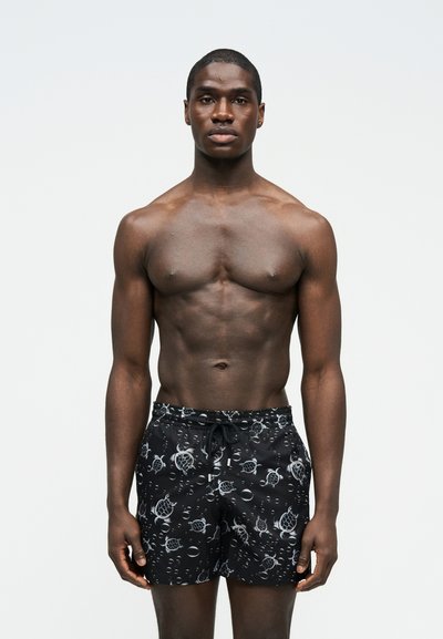 Μυώδης μαύρος άνδρας στέκεται μπροστά σε απλό φόντο, φορώντας μαύρο μαγιό με λευκά σχέδια θαλάσσιας χελώνας και φυσαλίδων.