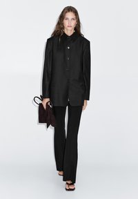 Black tailored blazer with a collared neckline, button closure, and long sleeves. Paired with black fitted pants and a dark suede handbag.