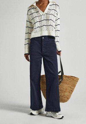 Sweter dziany w kolorze kremowym z granatowymi paskami, z dekoltem w kształcie litery V i szerokimi rękawami; zestawiony z ciemnoniebieskimi spodniami o szerokich nogawkach i plecioną torbą ze słomy.