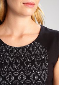 Svart topp med rund halsringning, strukturerad tyg med ett vitt geometriskt mönster och korta ärmar, som betonar kontrasterande färger.