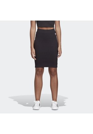 Vrouw die een zwarte aansluitende knie-lange rok en een cropped top draagt, gecombineerd met witte Adidas sneakers met zwarte strepen, staand op een effen achtergrond.
