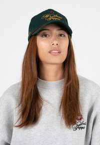 Groene pet met geborduurd gele tekst "De Zwitserse Yuppie" en bordeauxrode accenten. Grijze sweatshirt met logodetail en geribde kraag.