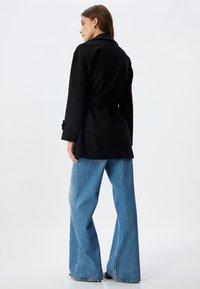 Sort belted frakke medindsnit, parret med lyseblå brede jeans. Frakken har en struktureret overflade og dekorative manchetter.
