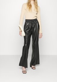 Kvinna i beige långärmad topp, svarta vida fusk läderbyxor, svarta öppna klackar och bär en liten vit flätad handväska.