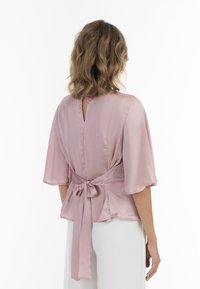 Rosenpink Bluse mit weiten Ärmeln, plissierter Textur, offener Rückseite mit Schlüsselöffnung und einer Schleifen-Detailung an der Taille.
