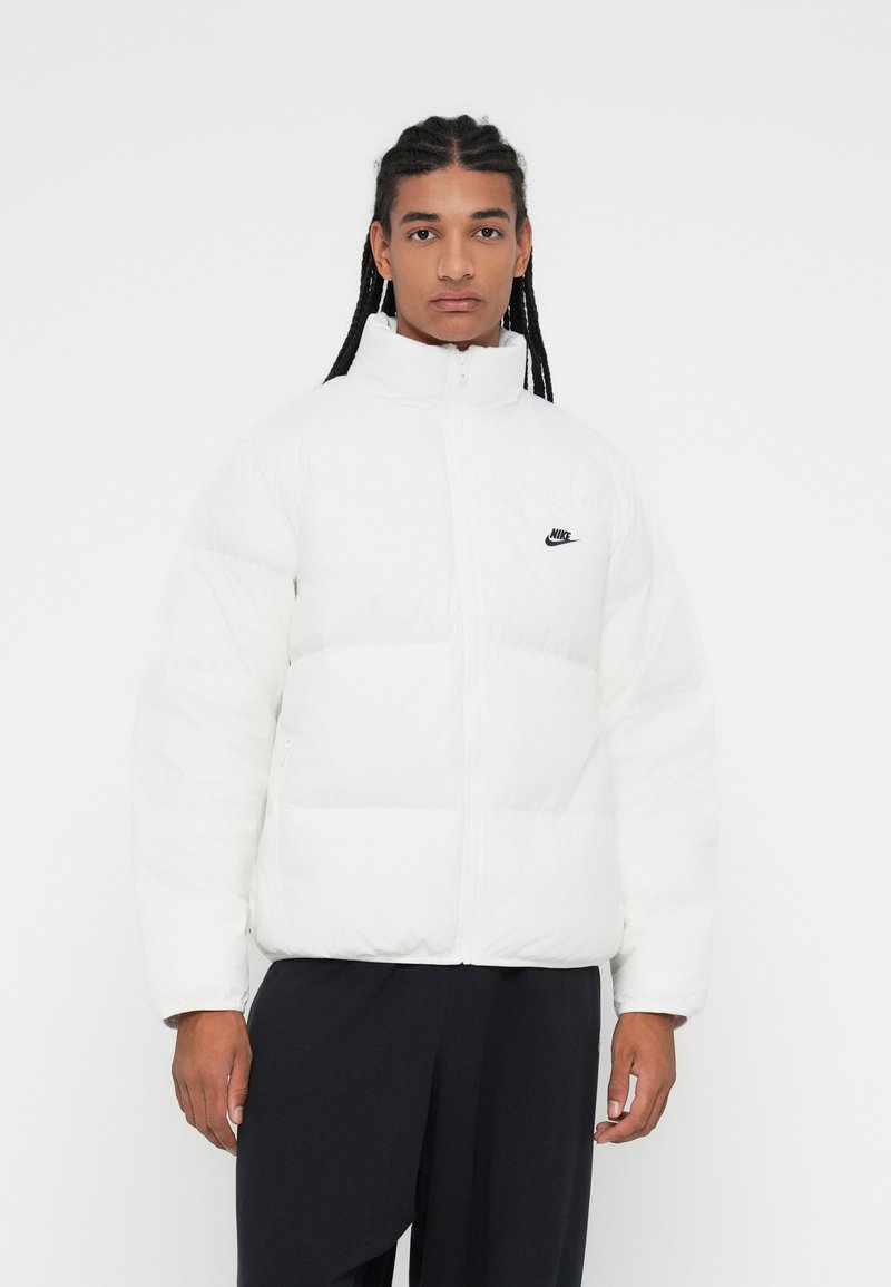 Bijela puffer jakna s visokim ovratnikom, zatvaranjem na patentni zatvarač i crnim Nike logotipom. Ima kviltani dizajn i meku, glatku teksturu.