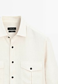 Linen shirt in a light beige color, featuring a button-down collar, black buttons, and two chest pockets with flaps.