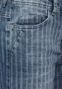 Vaqueros de mezclilla azul claro con desteñido vertical, un bolsillo delantero y detalle de botón metálico. Presentan una textura a rayas sutil y acentos de costura.