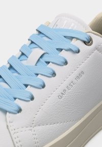 Baskets en cuir blanc avec des lacets bleu clair, une semelle beige, et l'inscription "GAP EST. 1969" embossée sur le côté.