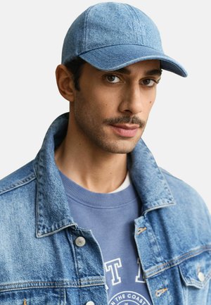 Uomo che indossa una giacca di jeans azzurro chiaro, un berretto di jeans abbinato e una felpa blu con testo bianco su sfondo bianco.
