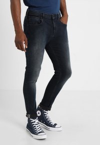 Dunkelblaue Jeans mit einem taillierten Design, ausgestattet mit fünf Taschen und einem Reißverschluss. Kombiniert mit navyfarbenen High-Top-Sneakern.