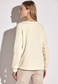 Světle béžová mikina s dlouhými rukávy, žebrovanými manžetami a širokým výstřihem, doplněná světle béžovými kalhotami. Hladká textura látky.