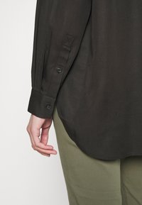 Dark green long-sleeved shirt with a curved hem, button cuffs, and a smooth texture, displayed against a light background.